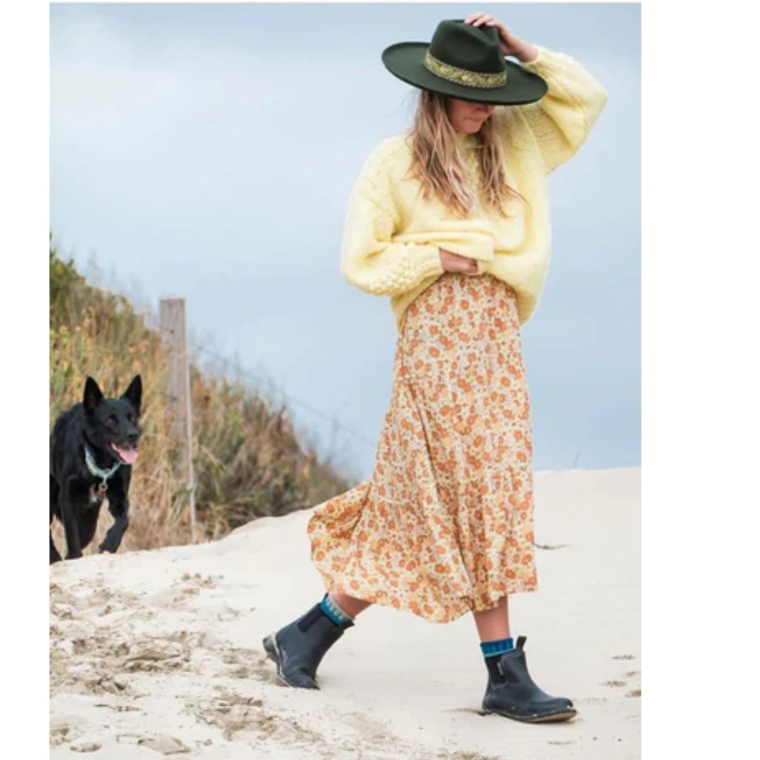 mulher andando na areia usando suéter amarelo, saia florida e bota preta