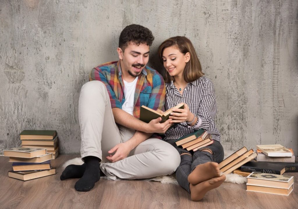 Casal sentado no chão lendo um livro juntos. Do lado deles estão vários livros empilhados.