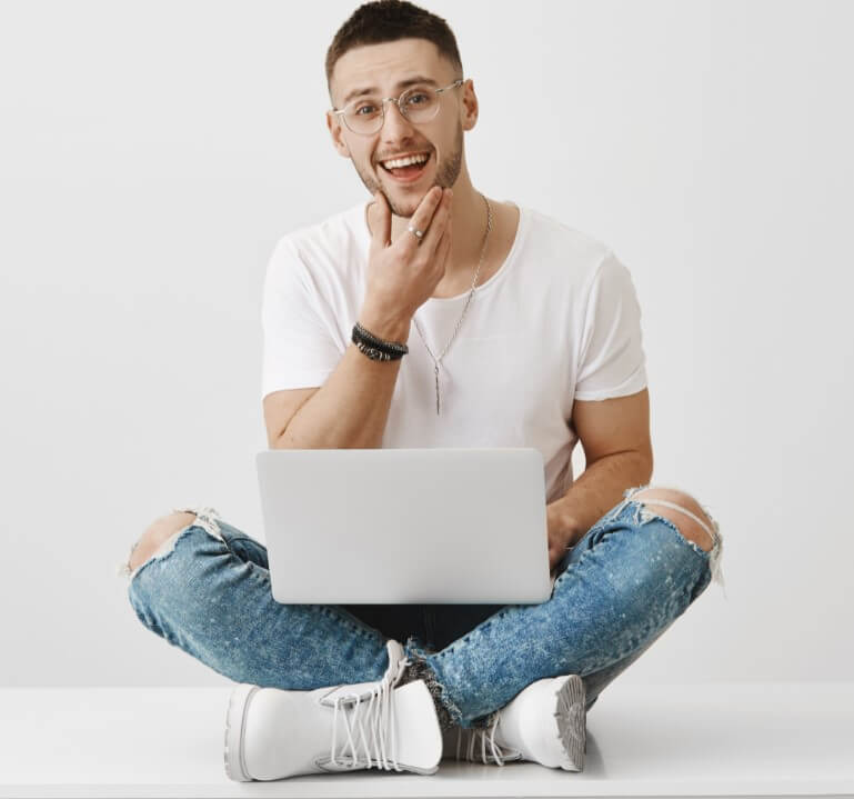 homem sentado no chão, com as pernas cruzadas, segurando um notebook no colo. Está sorrindo, passando a mão no queixo.