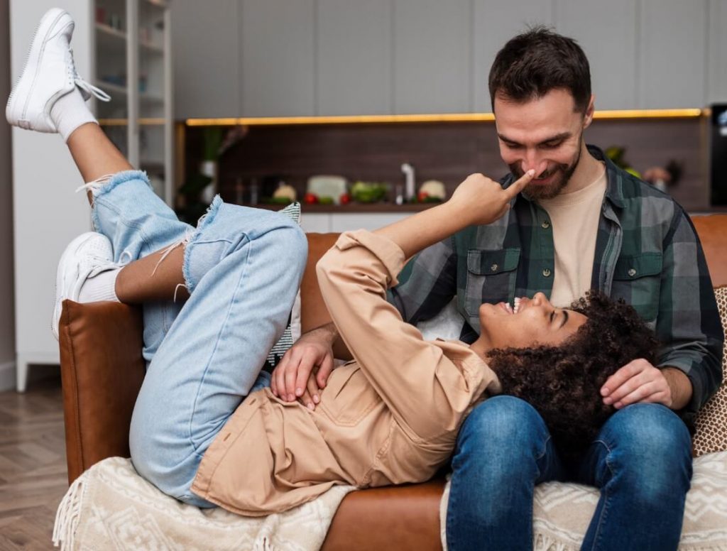 Casal na sala. O rapaz está sentado no sofá e a moça deitada com a cabeça no colo dele.