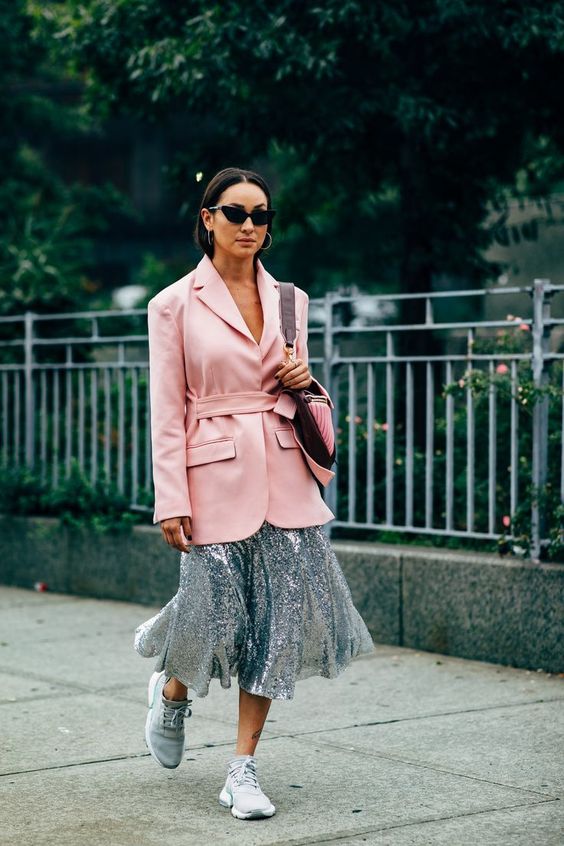 mulher andando pela rua com tênis cinza, saia cinza brilhante e maxi blazer rosa claro