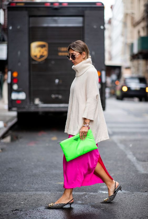 mulher atravessando a rua usa look de inverno com saia midi rosa neon, blusa bege de gola alta e bolsa verde neon