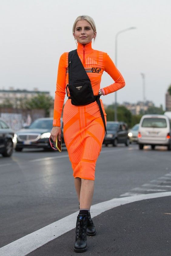 mulher jovem veste conjunto de saia e blusa laranja neon com bolsa preta transpassada no colo e coturno preto