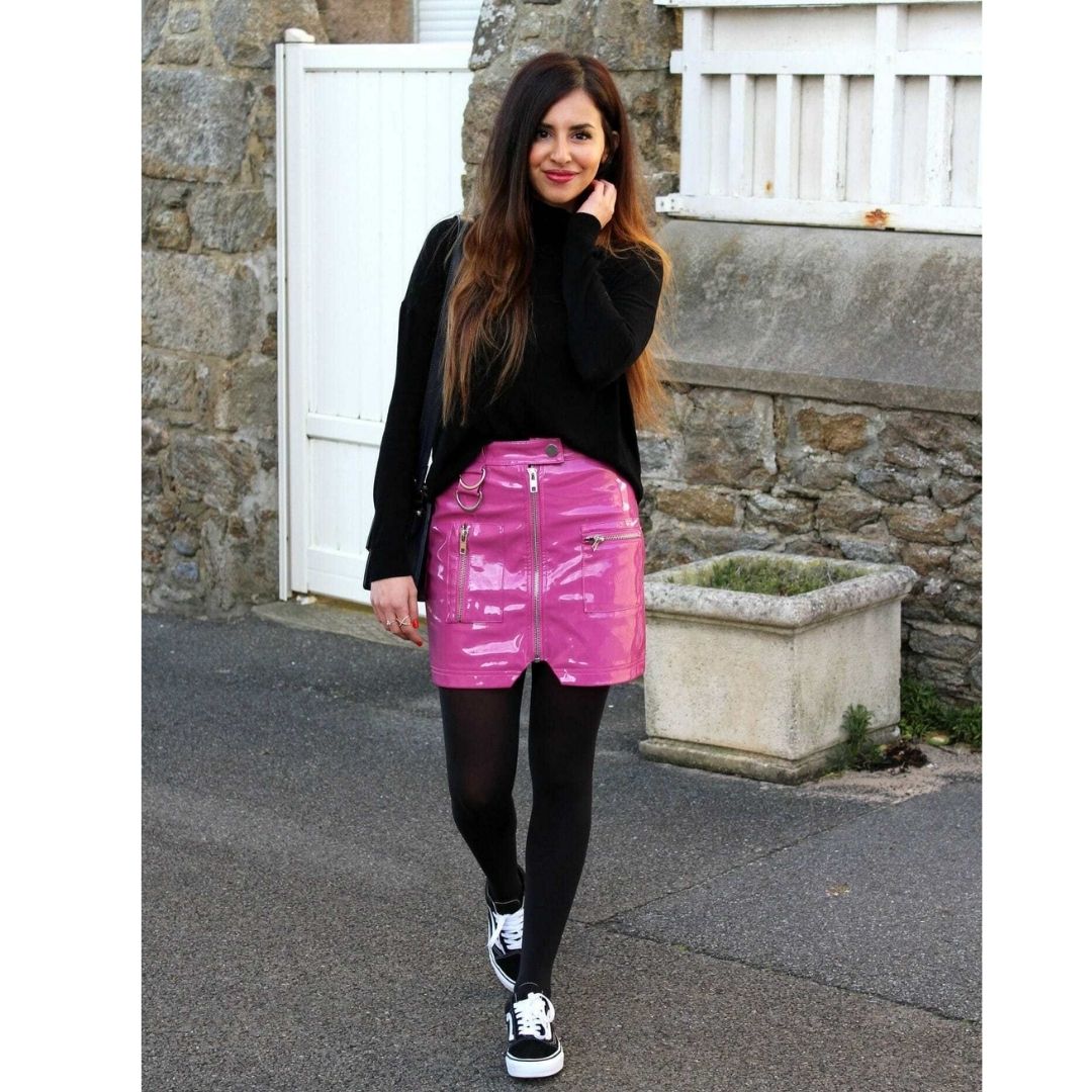 Mulher pousando para fotos na rua usando um look com tênis preto. meia calça, sai ade couro rosa e blusa preta