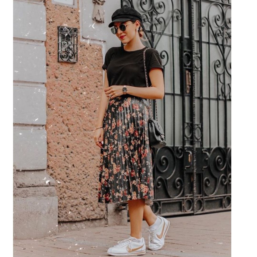 Mulher na rua com um look com tênis, sai midi floral, blusa preta, óculos de sol e uma boina 