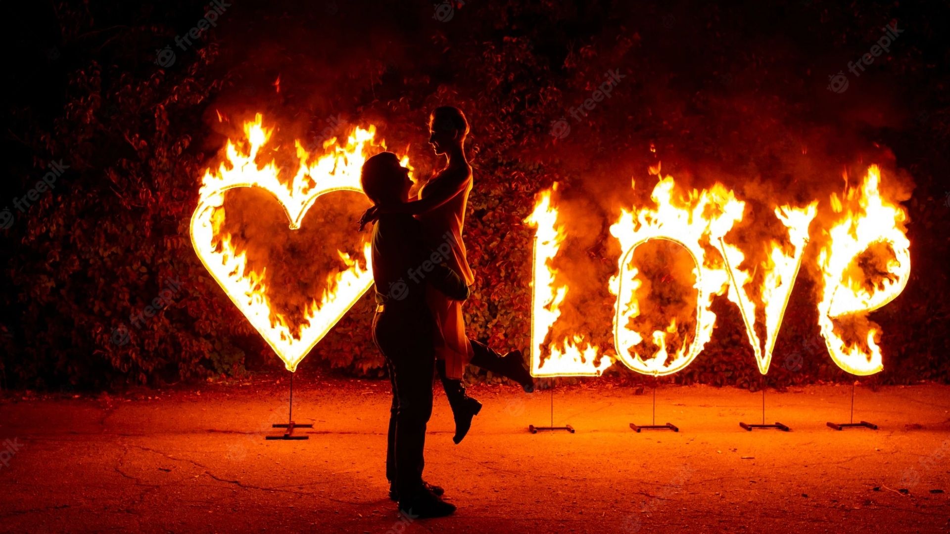Casal abraçados em frente um letreiro pegando fogo com a palavra love e um coração
