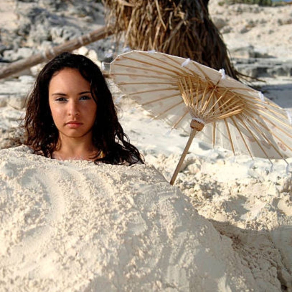 Mulher enterrada na areia com um guarda sol do lado, pousando para foto