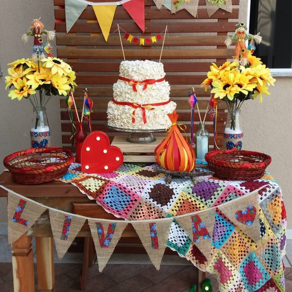 Mesa decorada com tema de festas juninas, bolo de pipoca, balão, flores e bandeirinhas.