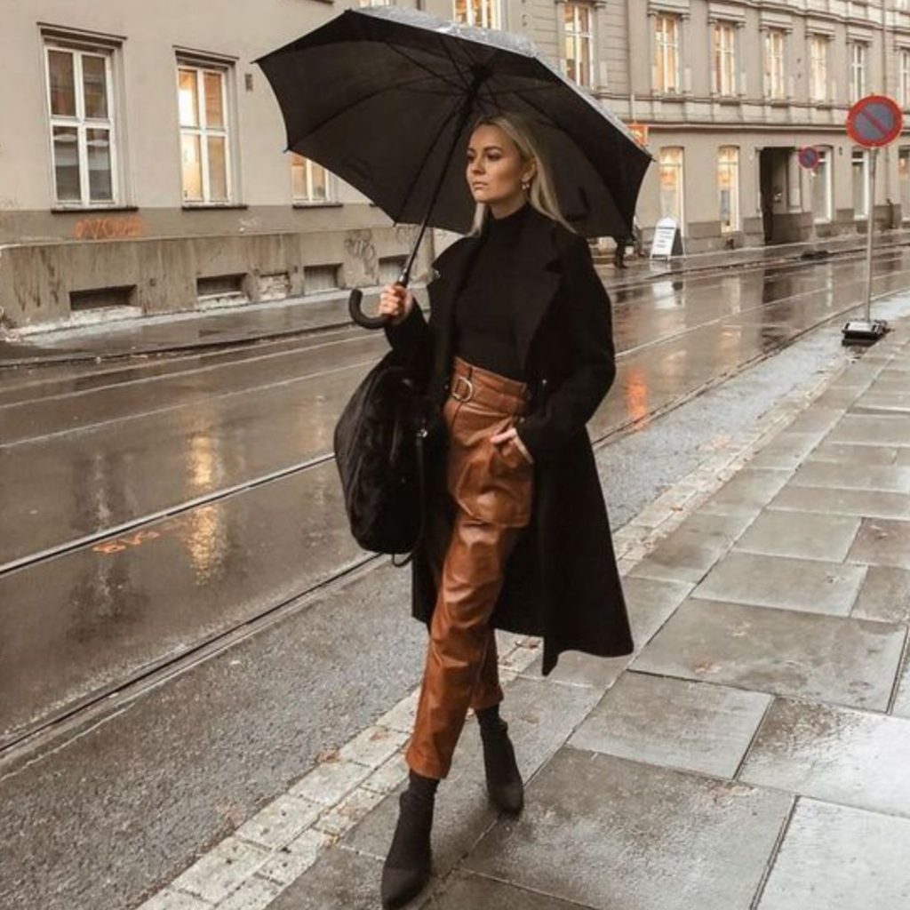 Mulher posando para fotos na rua usando uma calça de couro no tom mostarda, uma blusa preta, sobre tudo preto e um guarda chuva preto