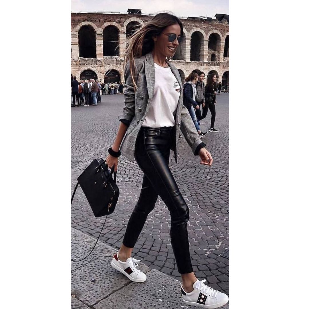 Mulher andando na rua com uma blusa branca, calça de couro preta, tênis branco, bolsa preta e casaco xadrez