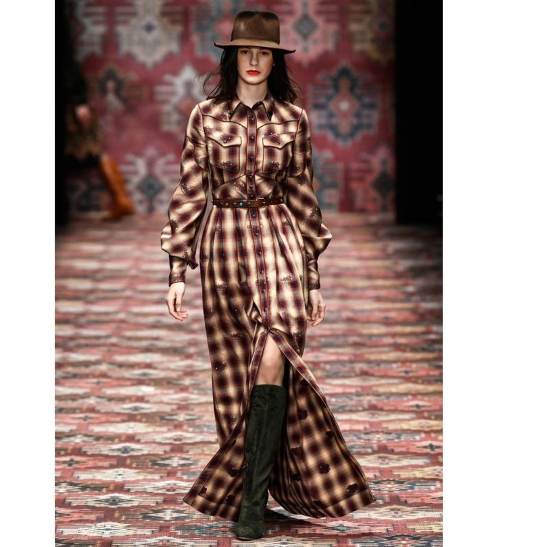Modelo desfilando look Western, composto por vestido longo xadrez, bota e chapéu.