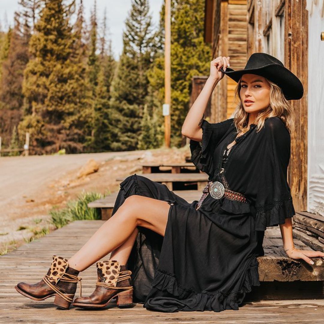 Modelo posando para foto com look Western, combinado vestido preto longo, chapéu, cinto e bota.