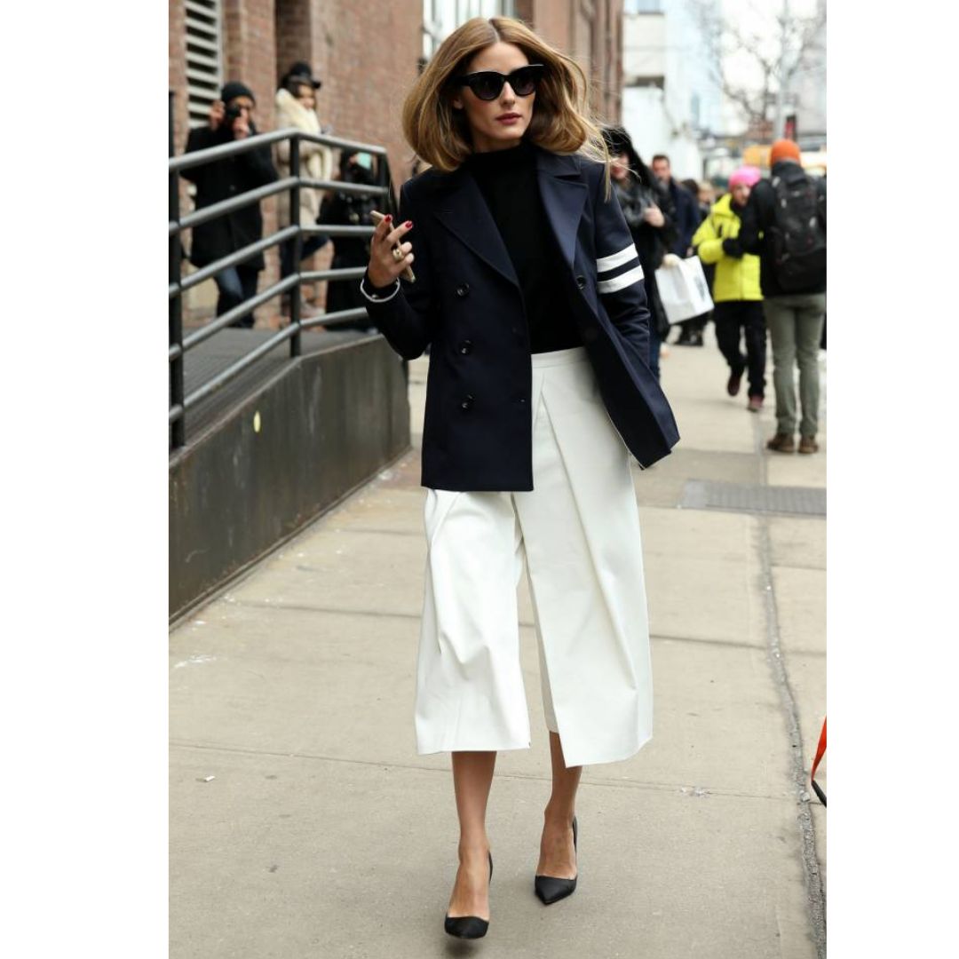 Mulher andando na rua vestindo uma calça pantacourt branca, blusa preta e uma jaqueta preta com detalhes em branco 