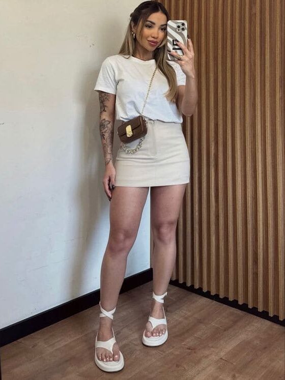 Mulher segurando celular para tirar foto do look com sandália para o verão. Chinelo flip flop branco, saia e camiseta branca.