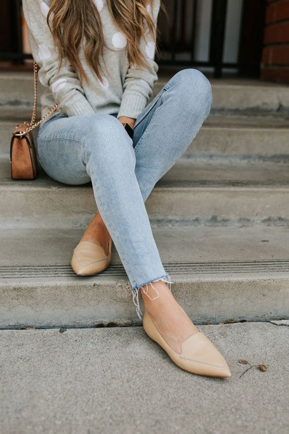 Mulher usando um look com calçado para inspirar você. O sapato é uma sapatilha de cor creme clara, jeans também claro e uma blusa de manga longa cinza. Ela também está usando uma bolsa pequena de cor castanha.