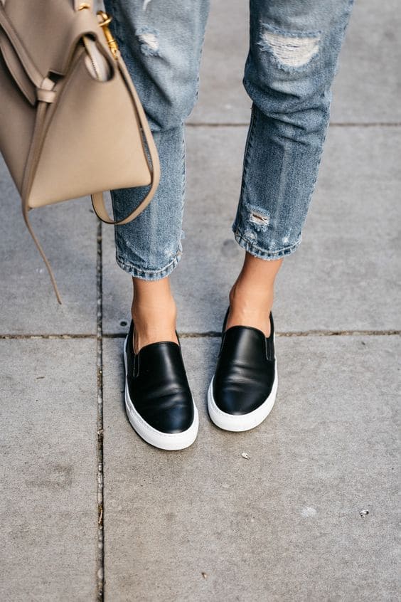Foto dos pés de uma mulher usando um calçado no estilo Slip-on, jeans com desfiados propositais e uma bolsa de cor creme.