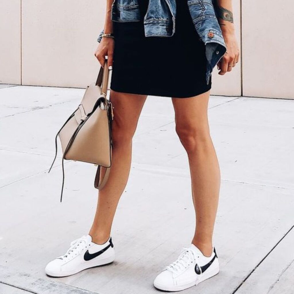 Mulher usando vestilo preto com uma jaqueta jeans presa à cintura, segurando uma bolsa de cor castanha e usando um tipo de tênis casual de cor branca da marca Nike