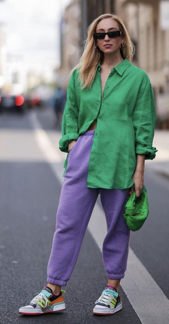 Mulher loira de cabelos lisos usando uma camisa grande de cor verde, calça de moletom de tom arroxeado e um tipo de tênis de skate.
