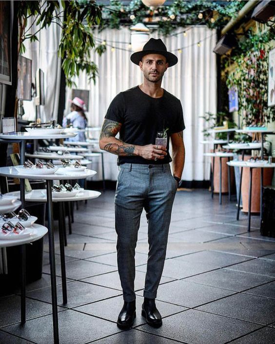 Homem usando um outfit mais formal com camisa e chepéu pretos, uma calça de alfaiataria na cor cinza e um tipo de sapato masculino mais social.