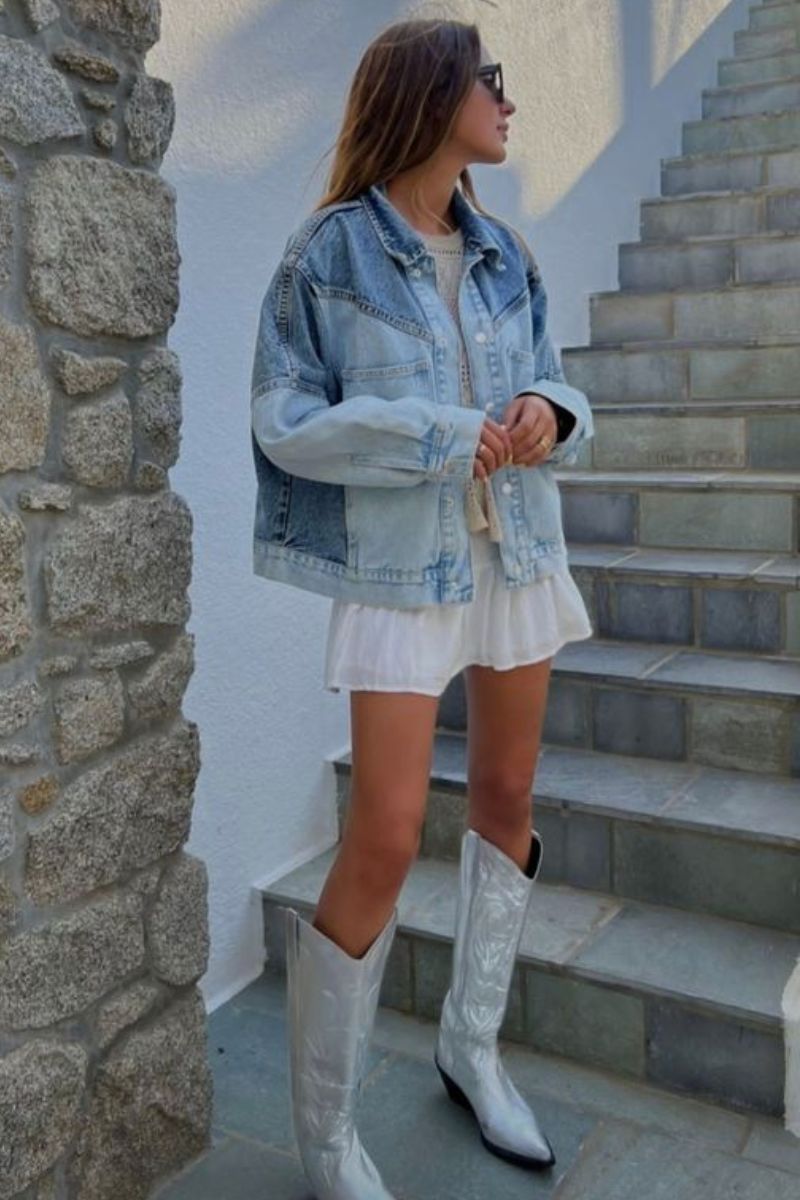 Mulher usando roupa que combina com bota composta por uma jaqueta jeans, vestido branco curto e bota de cano longo metalizada prata