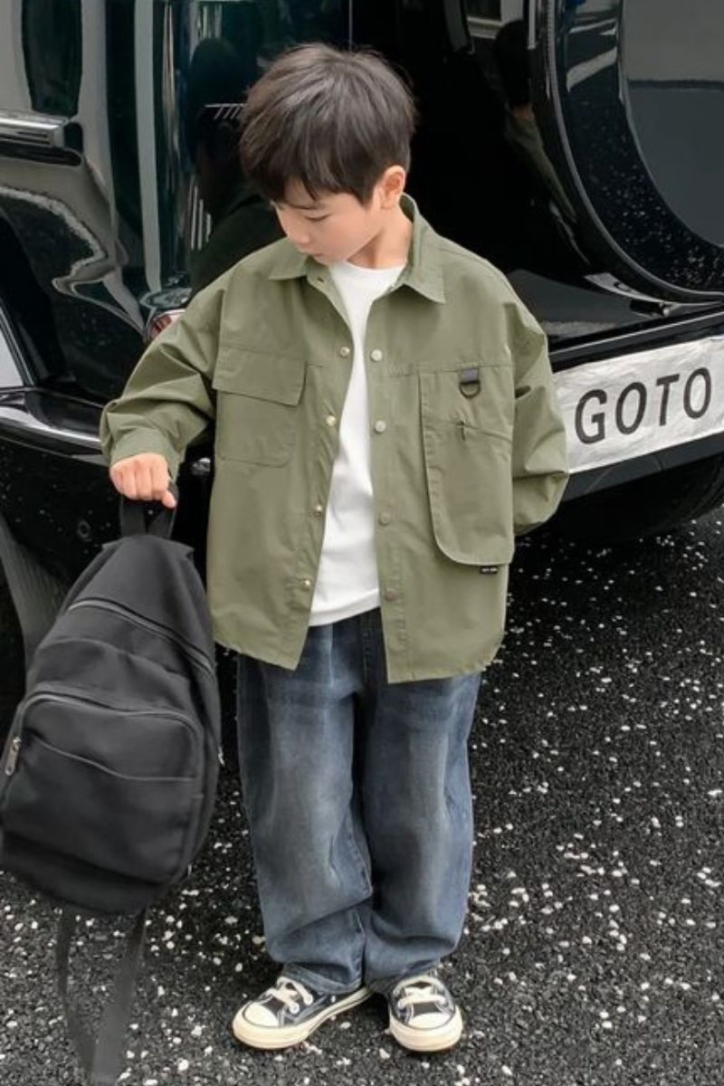 Menino asiático usando um look infantil com All Star de cor preta. Ele está segurando uma mochila com a sua mão direita.