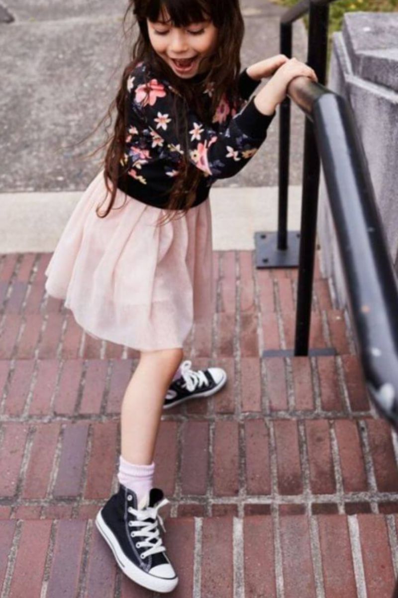 Menina pequena usando um look infantil com All Star composto por uma blusa de moleto preta florida e uma saia de pregas rosa