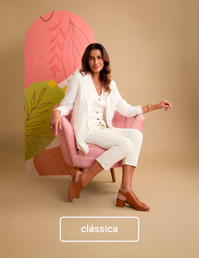 Mulher elegante com look branco e sandália marrom sentada em poltrona rosa, representando estilo clássico — inspiração de presente de Dia das Mães sofisticado e atemporal.