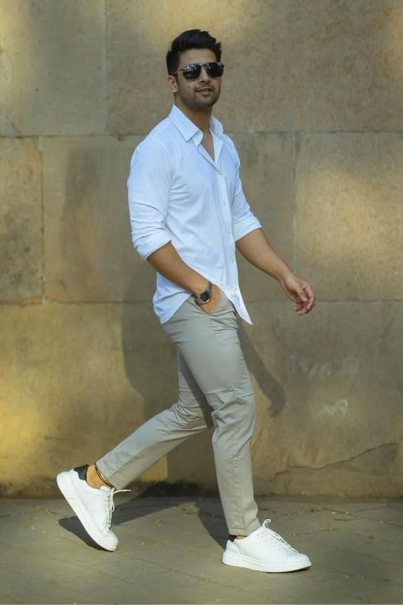 Homem moreno com barba por fazer e óculos escuros usando um look casual masculino composto por camisa e calça de cores claras.