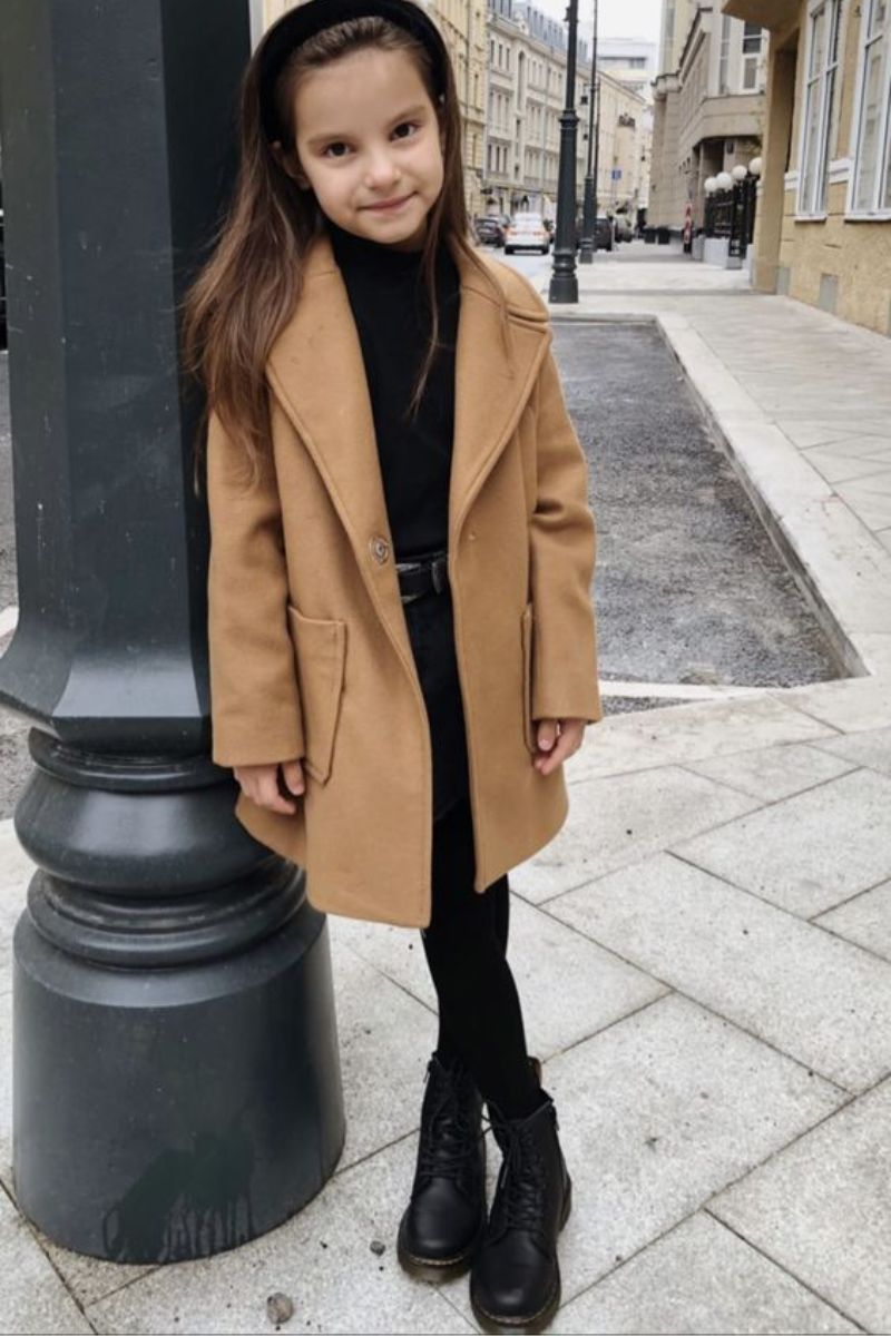 Menina pequena recostada num poste usando um look de inverno infantil feminino composto por sobretudo caramelo, outfit all black por baixo e bota