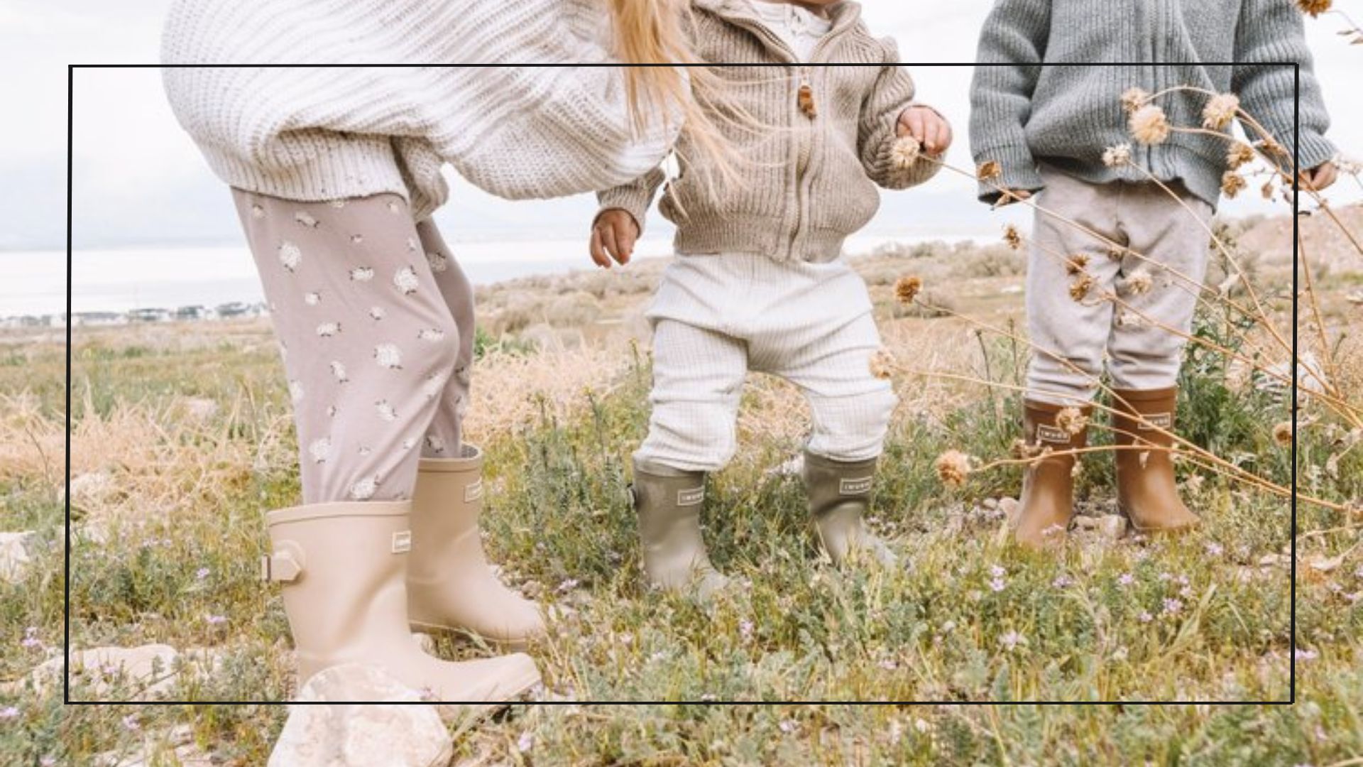 Três crianças usando looks infantis com bota brincando num campo com plantinhas