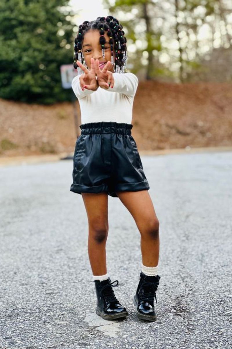 Menina negra usando um look infantil com bota composto por short de couro, blusa branca e coturno preto