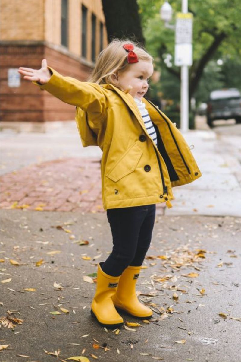 Menina pequena loira usando um look infantil com bota composto por galochas amarelas como sua jaqueta