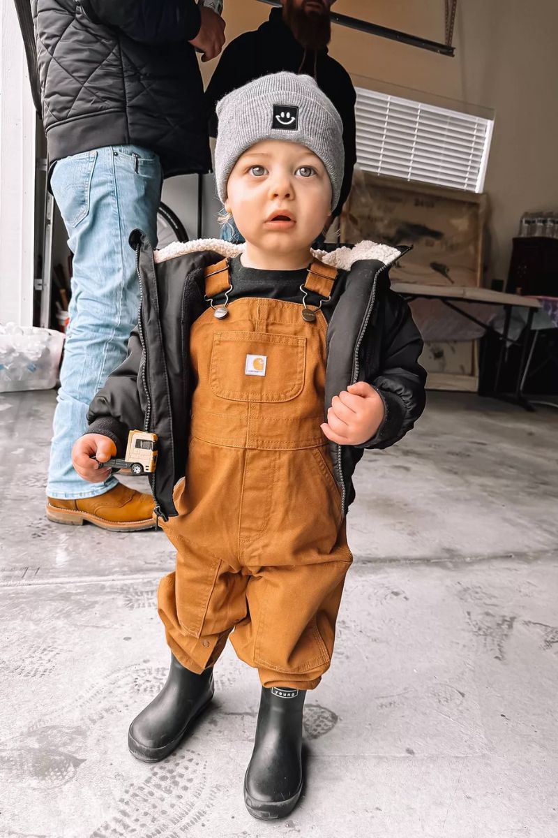 Menino pequeno usando um look infantil com bota composto por macacão de cor castanha e galocha preta