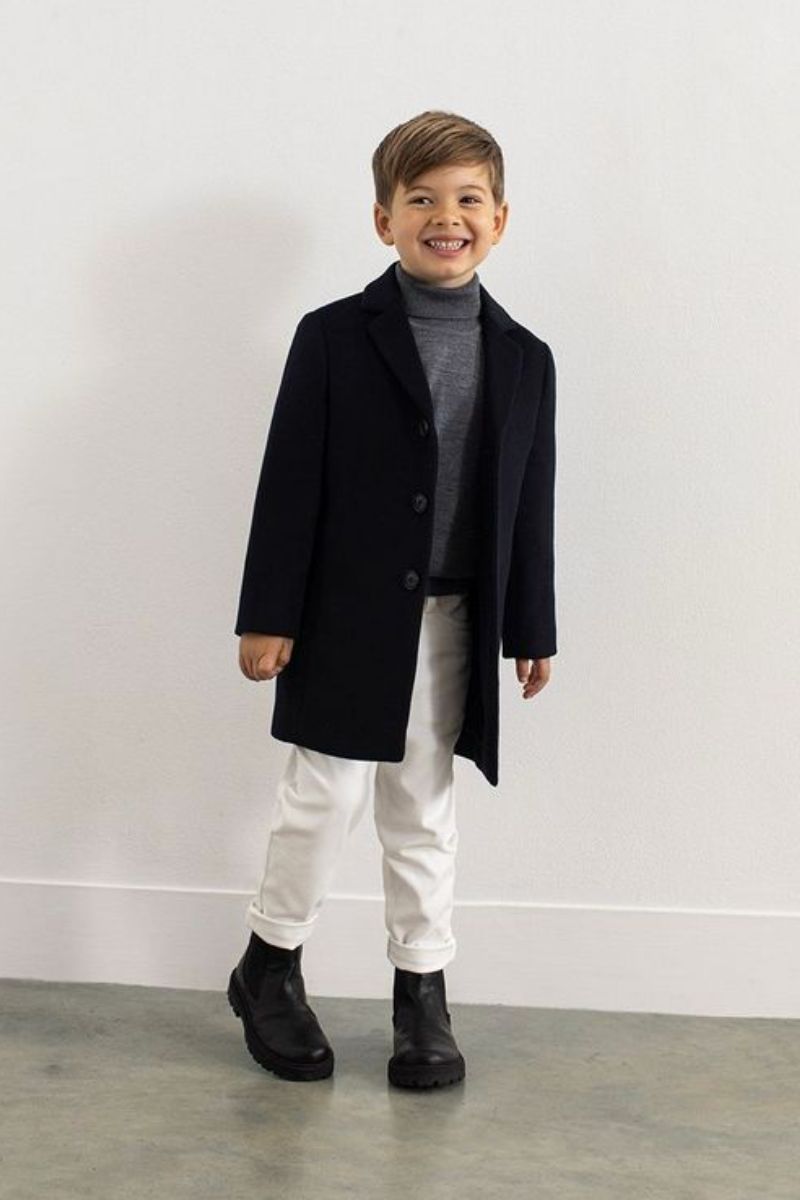 Menino de cabelos claros e lisos usando um look infantil com bota composto por bota Chelsea e sobretudo pretos