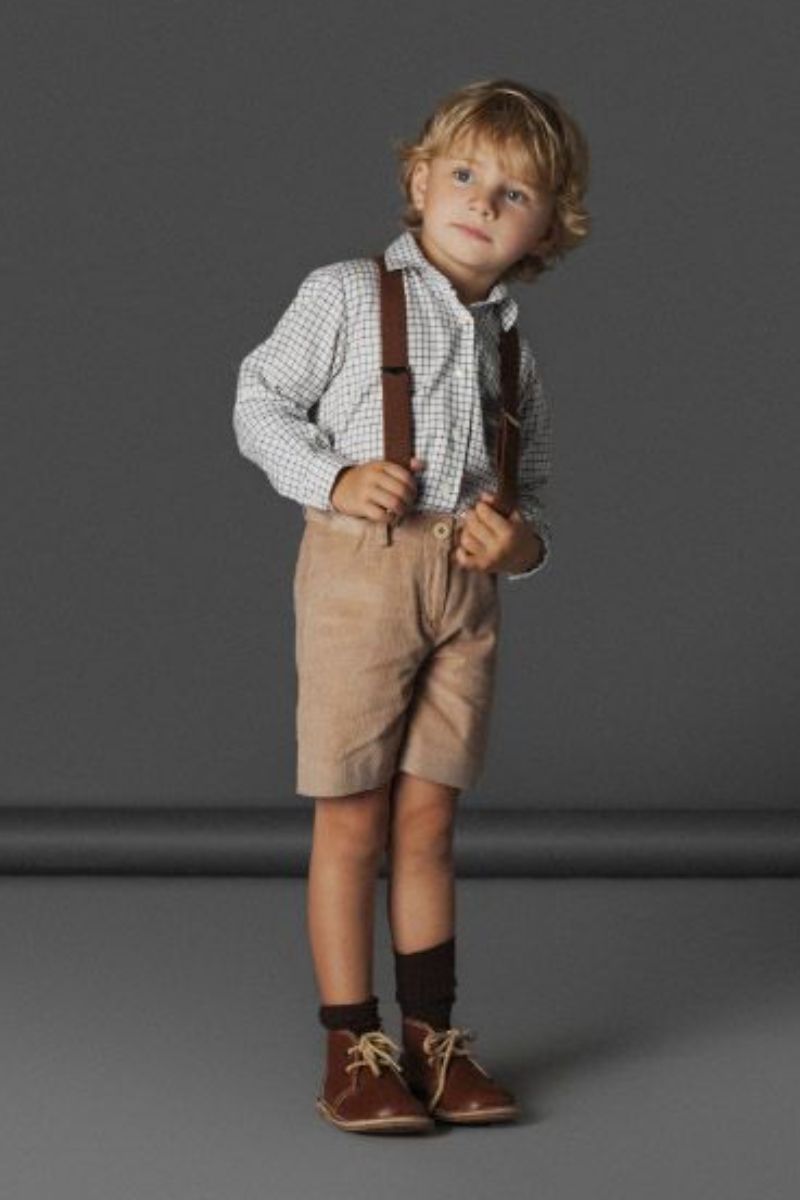 Menino loiro pequeno de olhos claros usando um look infantil com bota composto por bota marrom masculina de cano médio, camisa e suspensório