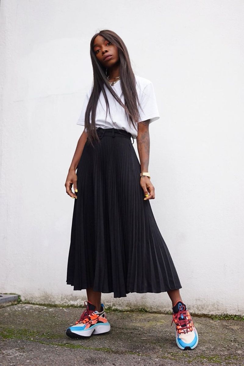 Mulher negra dando um exemplo do que é tênis chunky com saia longa preta e camiseta branca