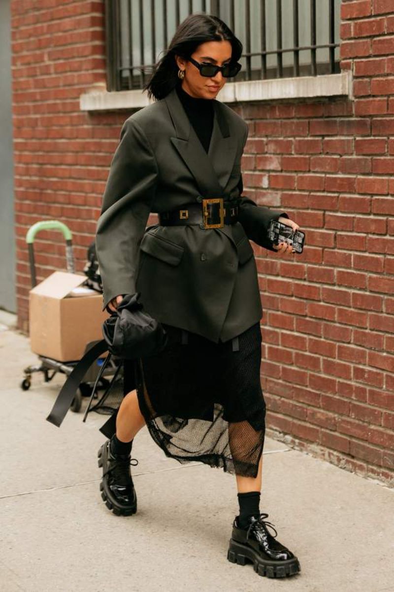 Mulher morena usando um look de inverno com saia e sapato preto e um blazer de alfaiataria chumbo