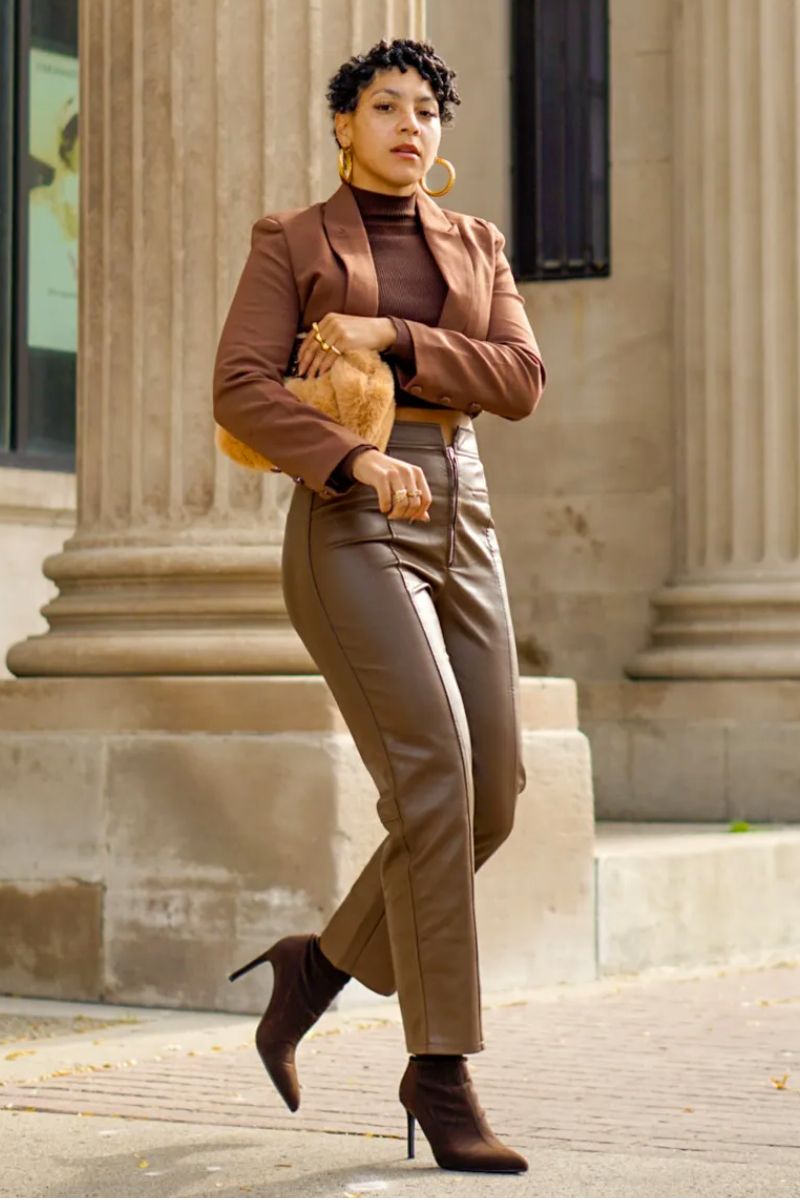 Mulher de cabelos curtos e morenos usando um look com bota cano curto marrom composto por calça de couro, blazer e blusa de gola alta.