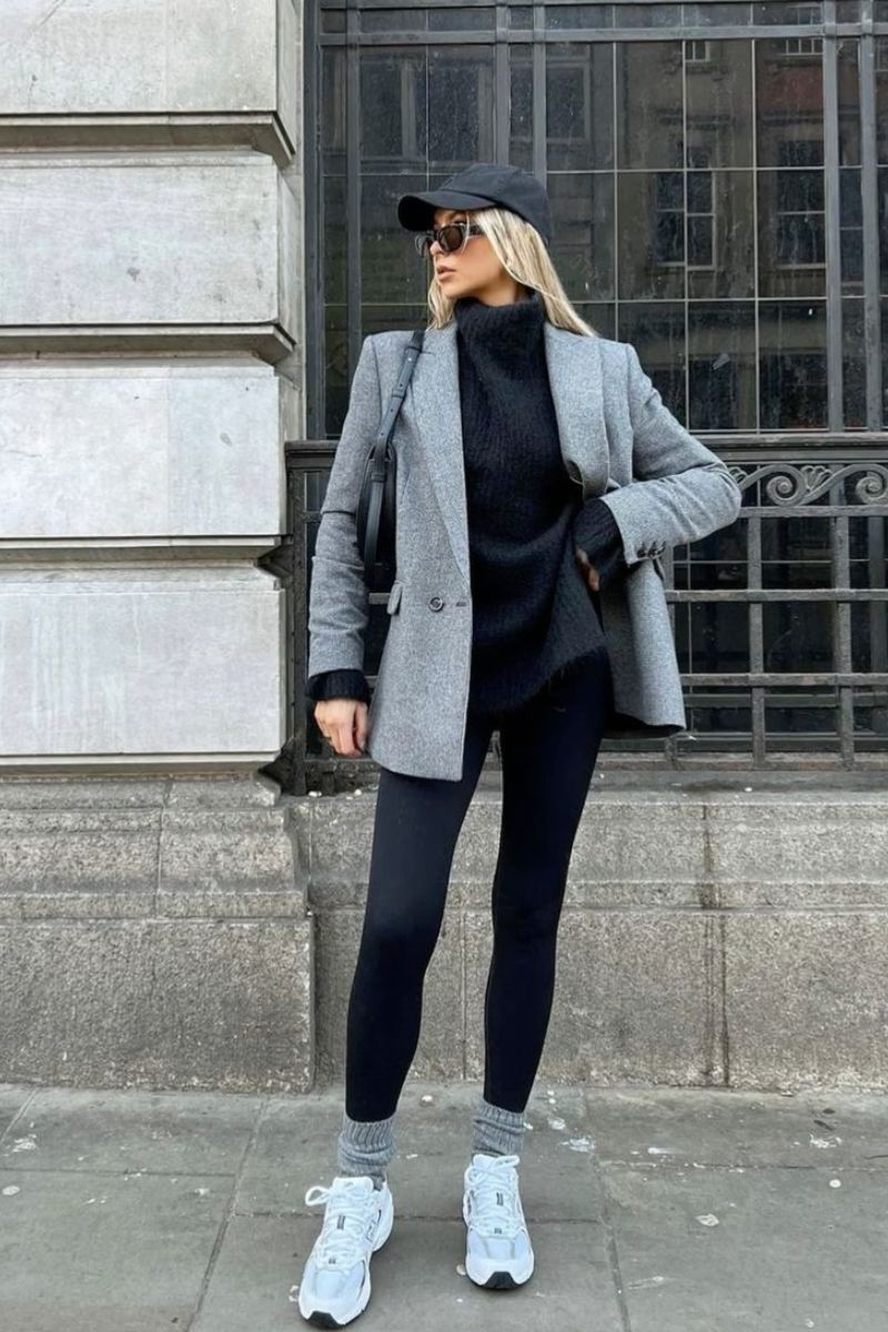 Mulher de cabelos loiros soltos usando um look predominantemente preto, com boné, blazer cinza e tênis confortável para trabalhar