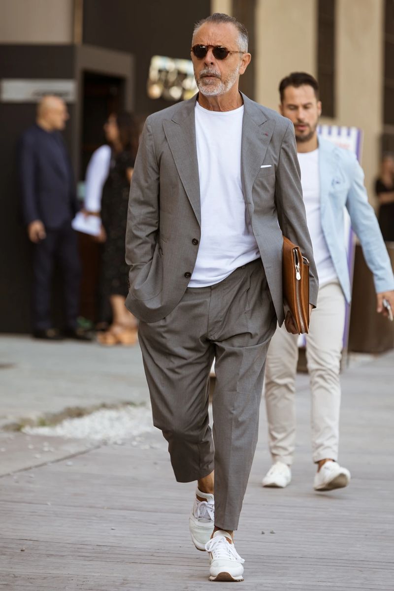 Homem de barba grisalha usando um look composto por peças de alfaiataria e tênis confortável para trabalhar de estilo esportivo de cor branca