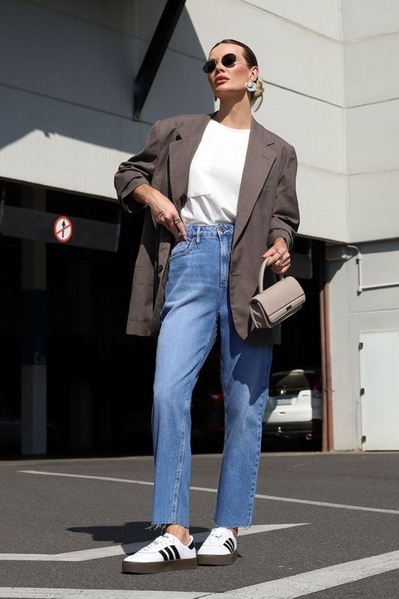 Mulher branca de cabelos presos e óculos escuros, camiseta branca, jeans, blazer e tênis confortável para trabalhar