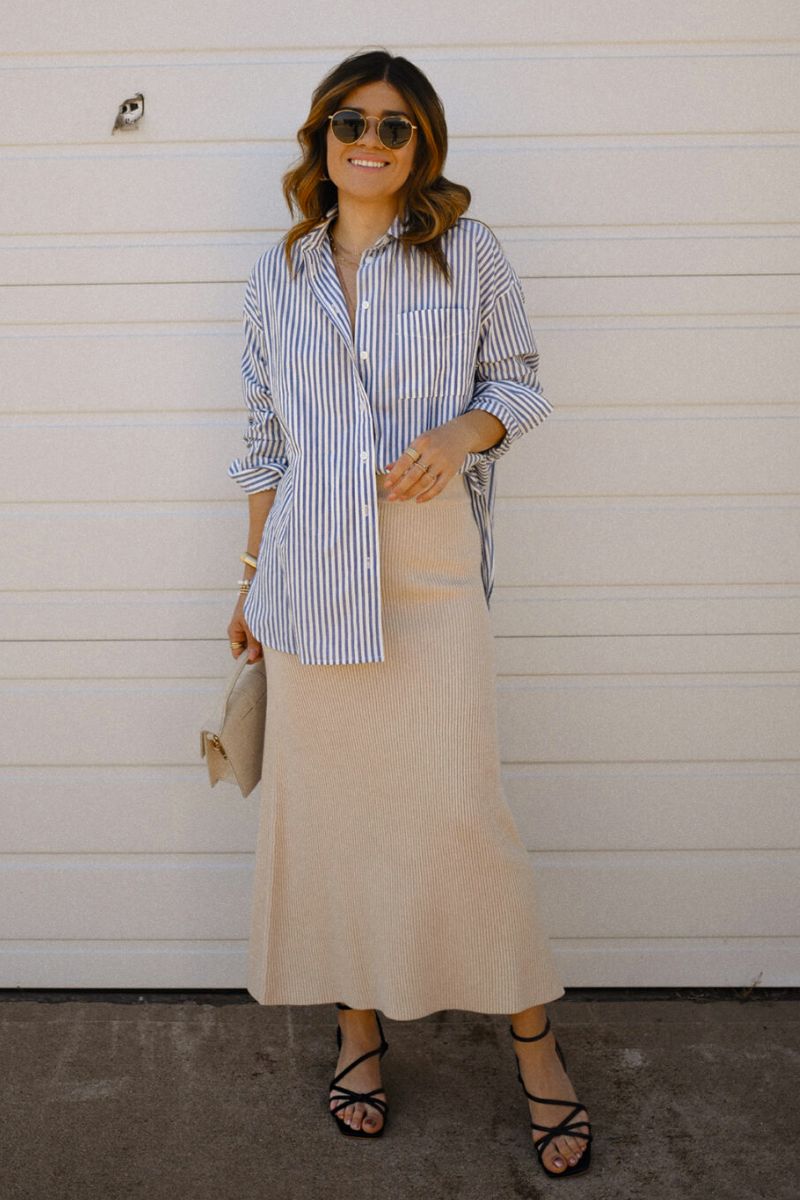 Mulher de óculos escuros usando um look com saia de tricot midi de cor areia e camisa de botão listrada azul e branca