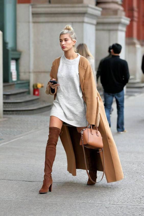 Mulher caminhando na cidade usando suéter oversized cinza como vestido, casaco longo caramelo e botas over the knee marrom de salto alto. Ela carrega uma bolsa estruturada em tom terroso e segura um celular.