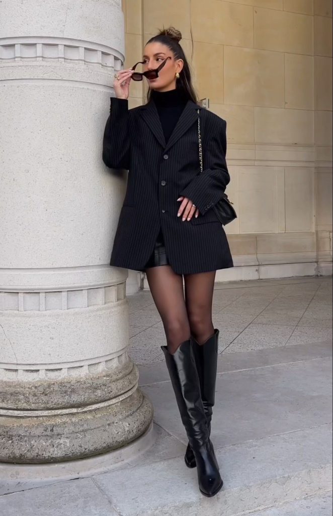 Mulher posando ao lado de uma coluna de pedra, vestindo blazer preto com risca de giz sobre blusa preta de gola alta, shorts curtos e meia-calça preta, combinados com bota de cano alto preta com salto. Ela segura óculos escuros e carrega bolsa preta com corrente dourada.